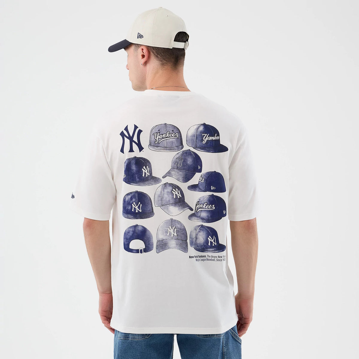 New Era New York Yankees MLB Cap Graphic White Oversized T-Shirt, back view on a model showcasing a large print of various blue and white Yankees caps, with the NY logo prominently displayed, and a New Era logo embroidered on the sleeve.