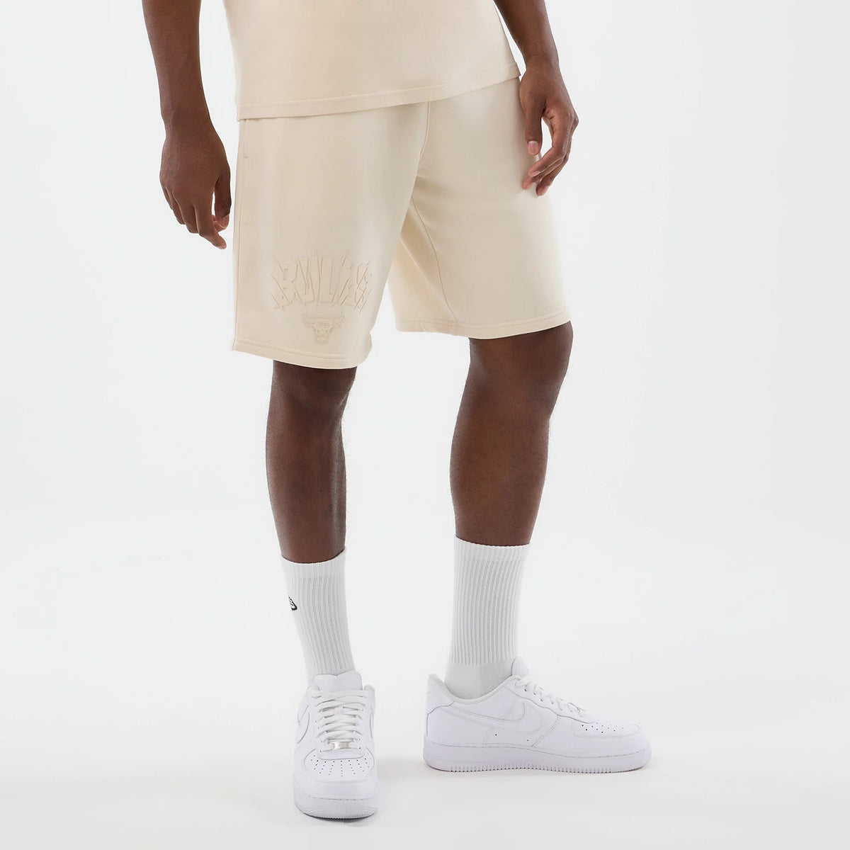 New Era Chicago Bulls NBA Core Plus Light Beige Shorts, front view on a model styled with white sneakers and socks, showing an embroidered Bulls logo and lettering across the upper left thigh, adding a team detail to the light beige fabric.