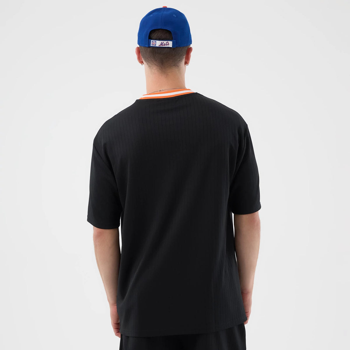 New Era New York Mets MLB Arch Black Oversized T-Shirt, back view on a model with a blue cap, showcasing the solid black back of the oversized shirt, a pop of orange around the neckline, and the cap featuring Mets lettering above the closure.
