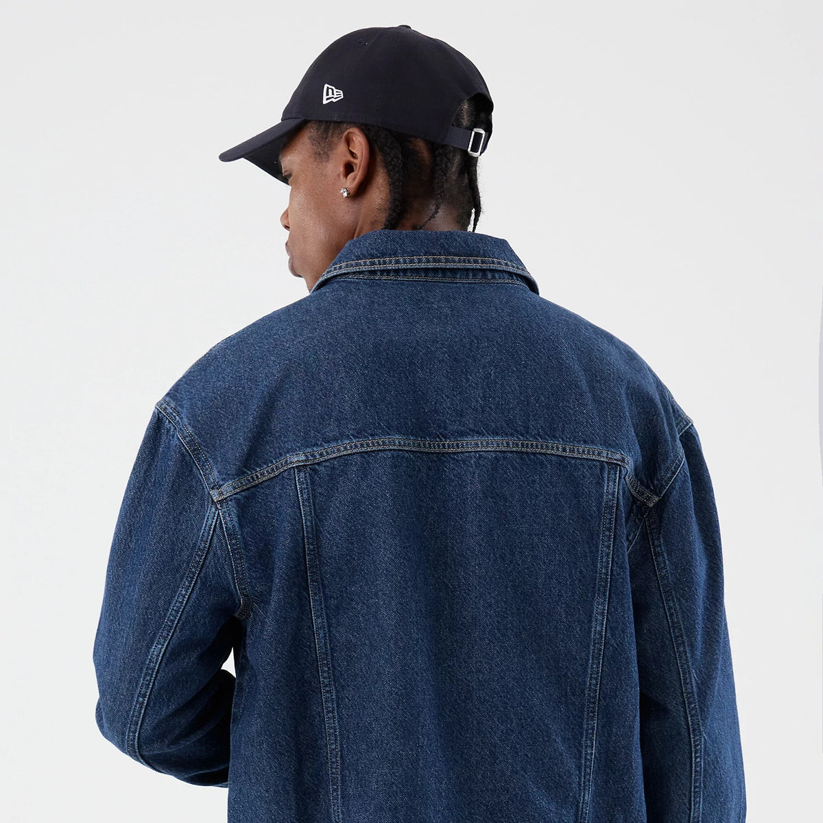 New Era Script Denim Dark Blue Jacket, back view on a model showing the rich indigo denim fabric, straight seam across the shoulders, and a classic fold-over collar accompanying a black New Era cap with a small embroidered logo.