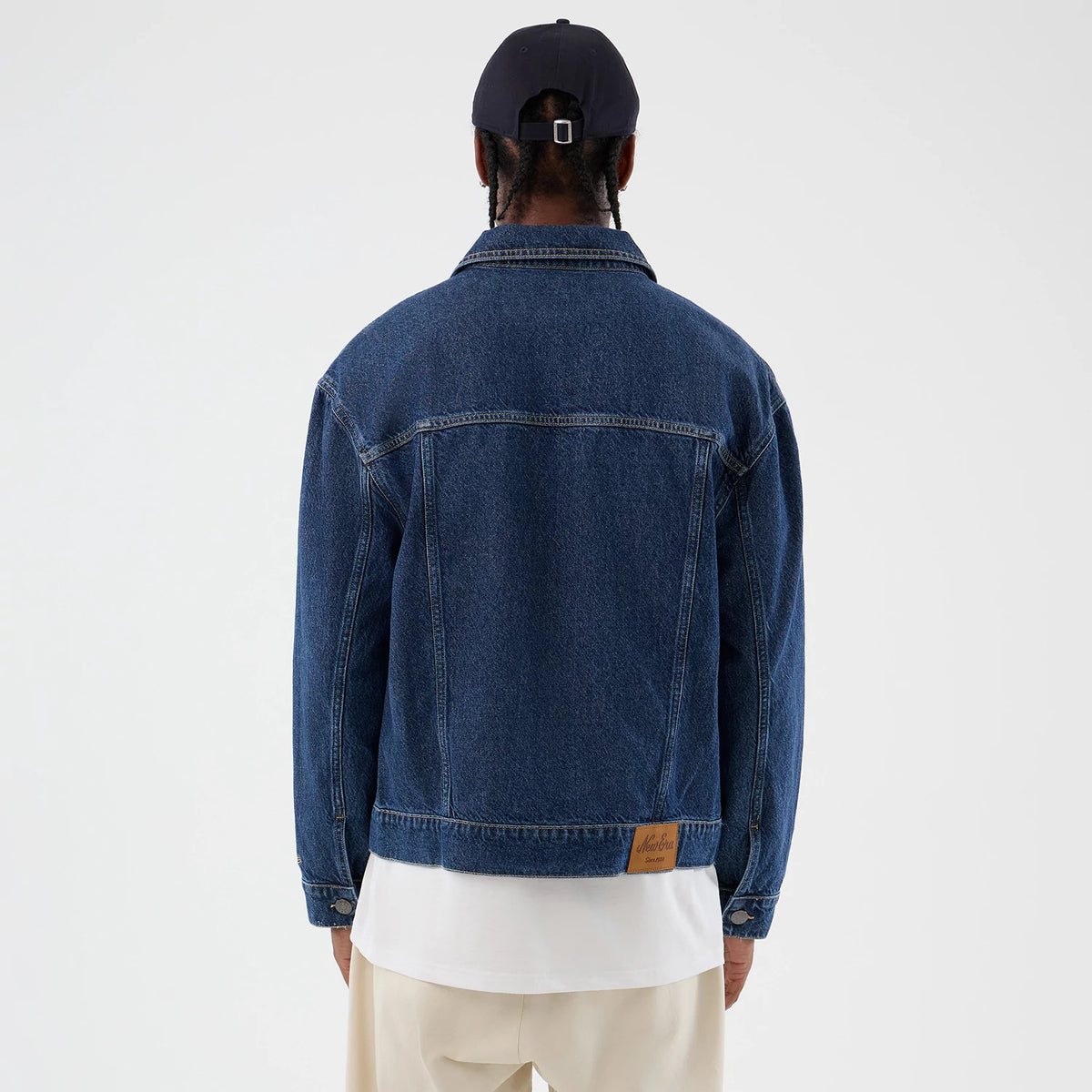 New Era Script Denim Dark Blue Jacket, back view on-model displaying the clean dark blue denim finish, pronounced seams running across the shoulders and back, and a leather patch with embossed script logo at the hem over a white T-shirt.