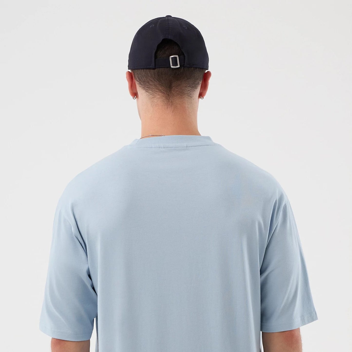 New Era New York Yankees MLB Script Pastel Blue Oversized T-Shirt, back view on a model with a navy cap, showing the plain pastel blue fabric with a loose fit and a neckline seam, complementing the simple and casual style.