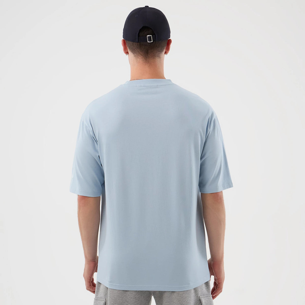 New Era New York Yankees MLB Script Pastel Blue Oversized T-Shirt, back view on a model wearing a dark cap, showing the simple pastel blue fabric with loose-fitting sleeves and a straight hem, paired with grey shorts for a casual look.