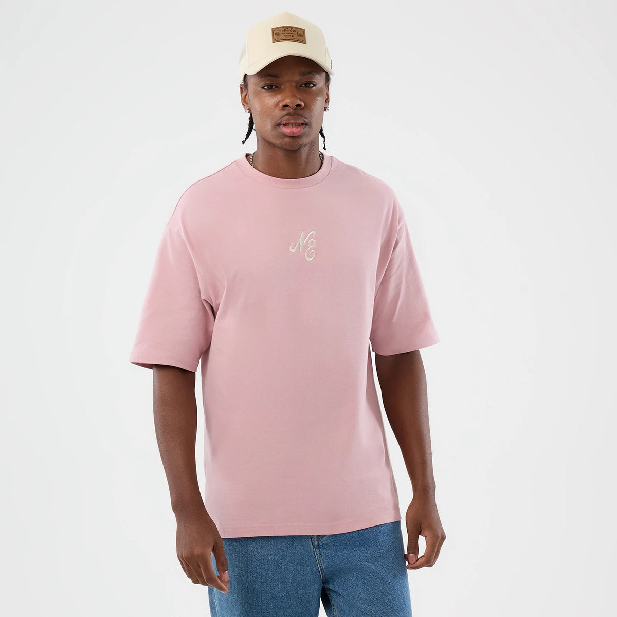 New Era Floral Graphic Pastel Pink Oversized T-Shirt, front view on a model with blue jeans and a cap, displaying the oversized pastel pink tee featuring an embroidered NE logo in white on the chest, offering a casual and look.