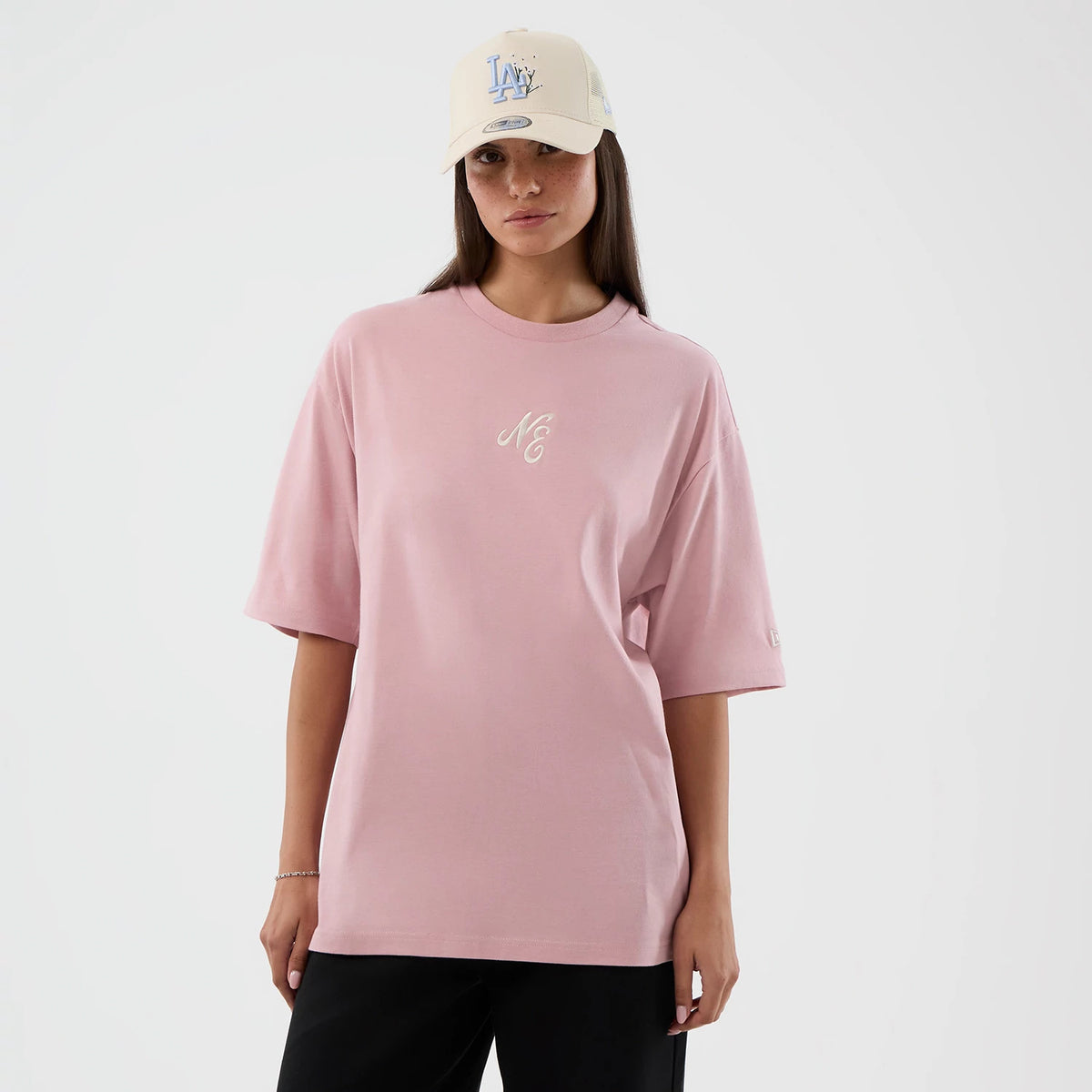 New Era Floral Graphic Pastel Pink Oversized T-Shirt, front view on-model showing embroidered NE logo on the chest, oversized short sleeves, and small detail on left shoulder, paired with a cream cap featuring an LA logo.
