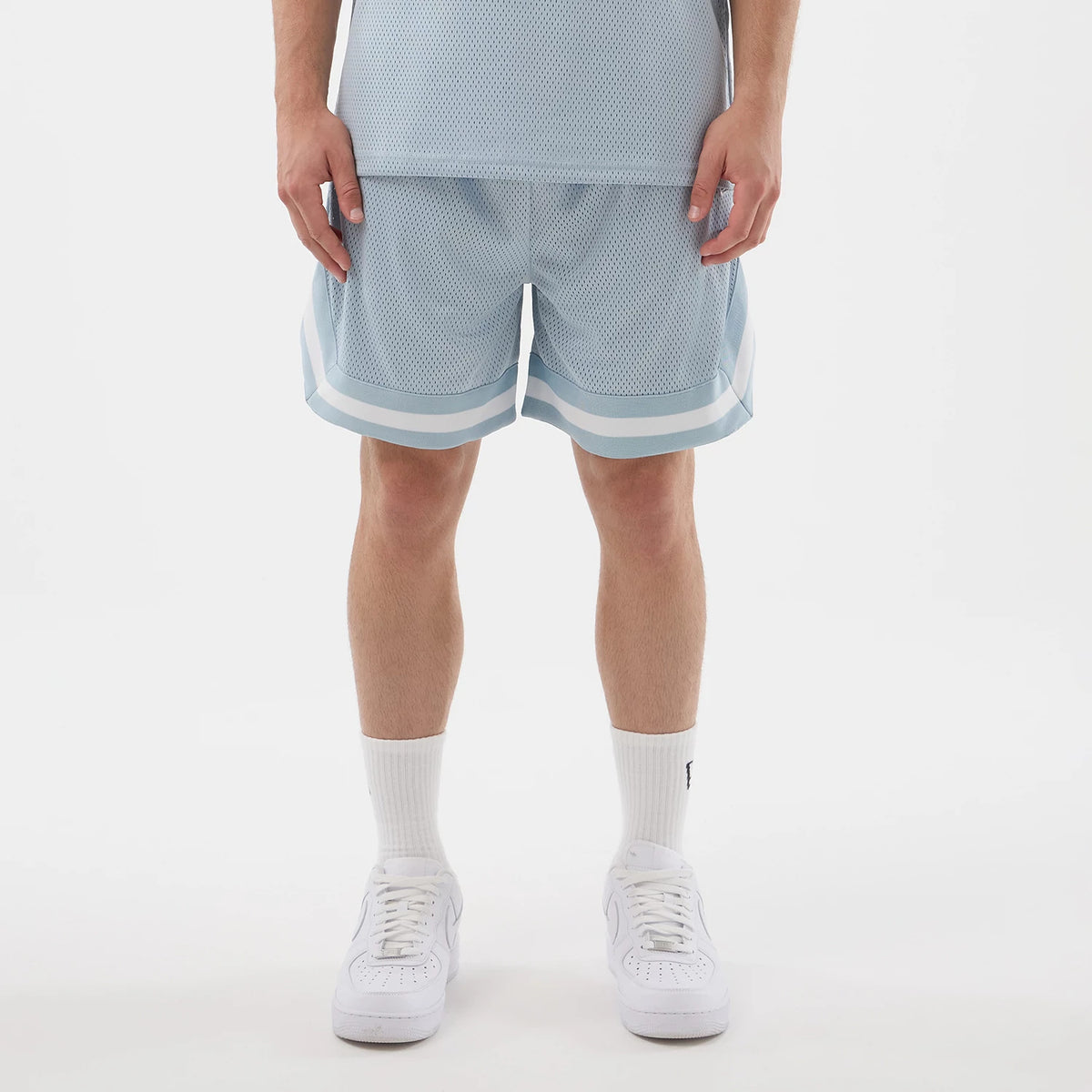 New Era Mesh Pastel Blue Oversized Shorts, front view on a model, showing light blue mesh fabric with white and light blue stripes along the hem, paired with white trainers and mid-calf white socks, creating a casual sporty look.