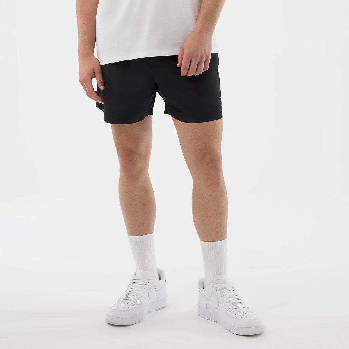 New Era Black Swimming Shorts, front view on-model showing the simple black design with a relaxed fit and above-the-knee length, styled with a white shirt and classic white trainers, giving a casual and appearance.