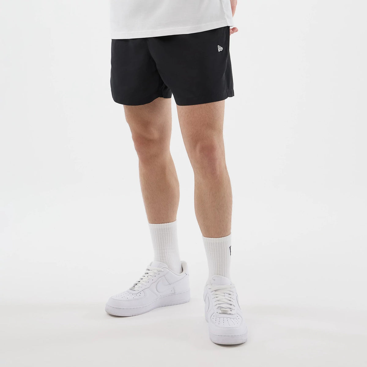 New Era Black Swimming Shorts, front view on a model paired with white sneakers and socks, showcasing the classic black shorts featuring a embroidered New Era logo in white on the left thigh, just above the hem.