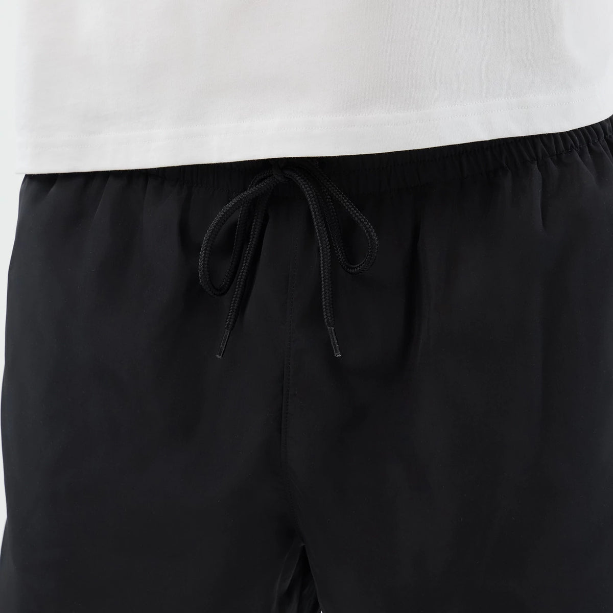 New Era Black Swimming Shorts, front view showing the plain black fabric with an elasticated waistband, paired with a white top. A black drawstring is tied at the centre of the waist, giving a casual, relaxed look to the shorts.