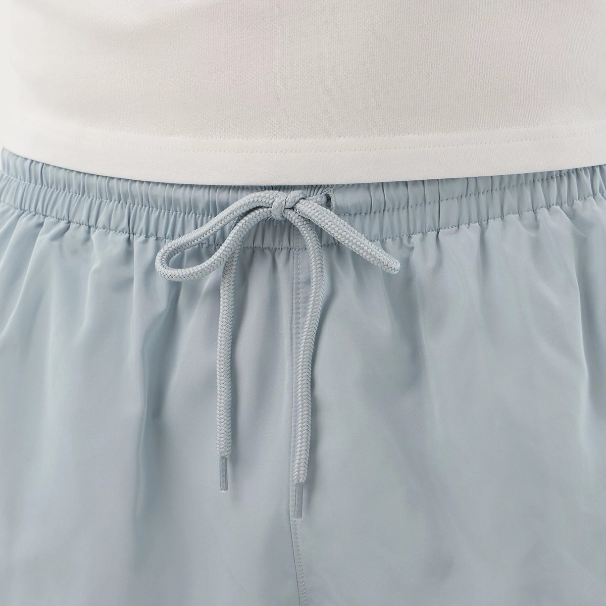 New Era Pastel Blue Swimming Shorts, front view showing the light blue fabric with an elasticated waistband, a drawstring tied in the centre, and the top edge of the paired white shirt just above the shorts.