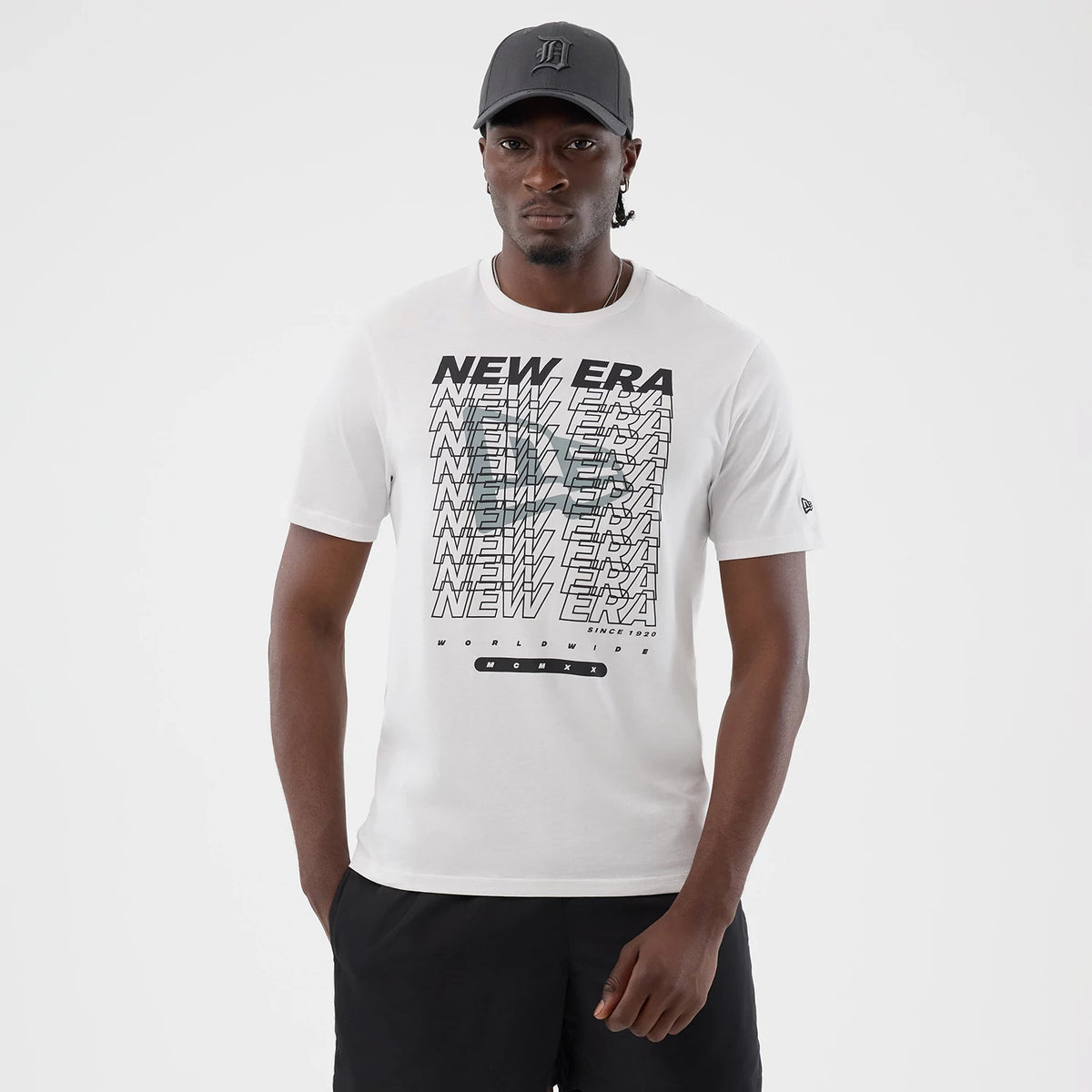 New Era Logo Graphic White T-Shirt, front view on a model wearing a dark cap and black shorts, featuring a graphic with multiple overlapping New Era texts across the chest and a small embroidered New Era flag logo on the upper left sleeve.
