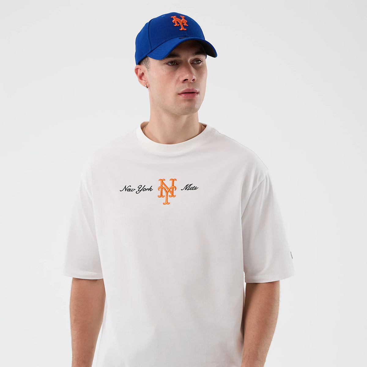 New Era New York Mets MLB Script White Oversized T-Shirt, front view on a model wearing a blue cap, showing orange and black embroidered New York Mets script and logo centred on the chest, with a minimal design and loose fit style.
