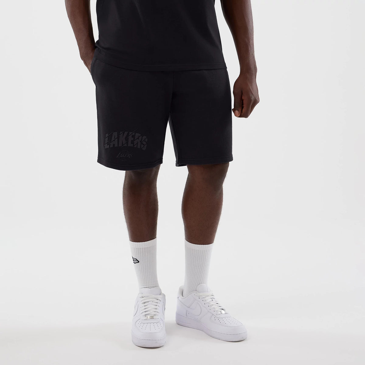 New Era LA Lakers NBA Core Plus Navy Shorts, front view on a model paired with a black top and white trainers, showing the word 'Lakers' in a graphic design printed on the left leg, with hands in pockets.