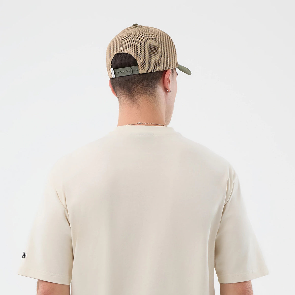 New Era Dessert Graphic Light Beige Oversized T-Shirt, back view on a model styled with a tan cap, displaying a plain light beige rear and an embroidered New Era logo on the left sleeve near the hem.