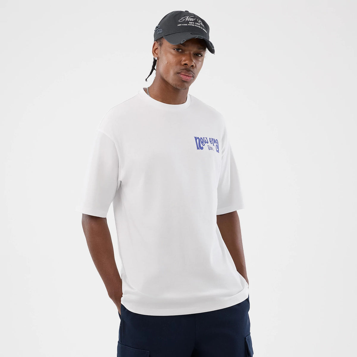 New Era Fun Pizza Graphic White Oversized T-Shirt, front view on a model teamed with a dark cap, featuring a blue printed New Era logo on the chest and short sleeves, creating a relaxed and casual style.