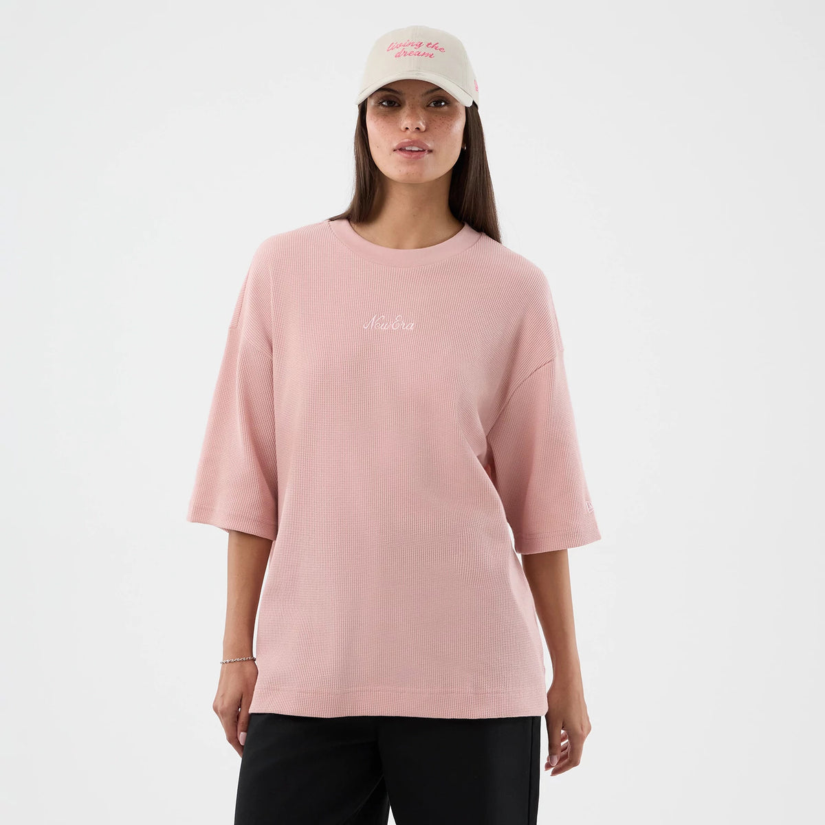 New Era Waffle Pastel Pink T-Shirt, front view on a model styled with black trousers and a cream cap, featuring a relaxed fit with half sleeves, a white embroidered New Era logo centred on the chest, and textured fabric.