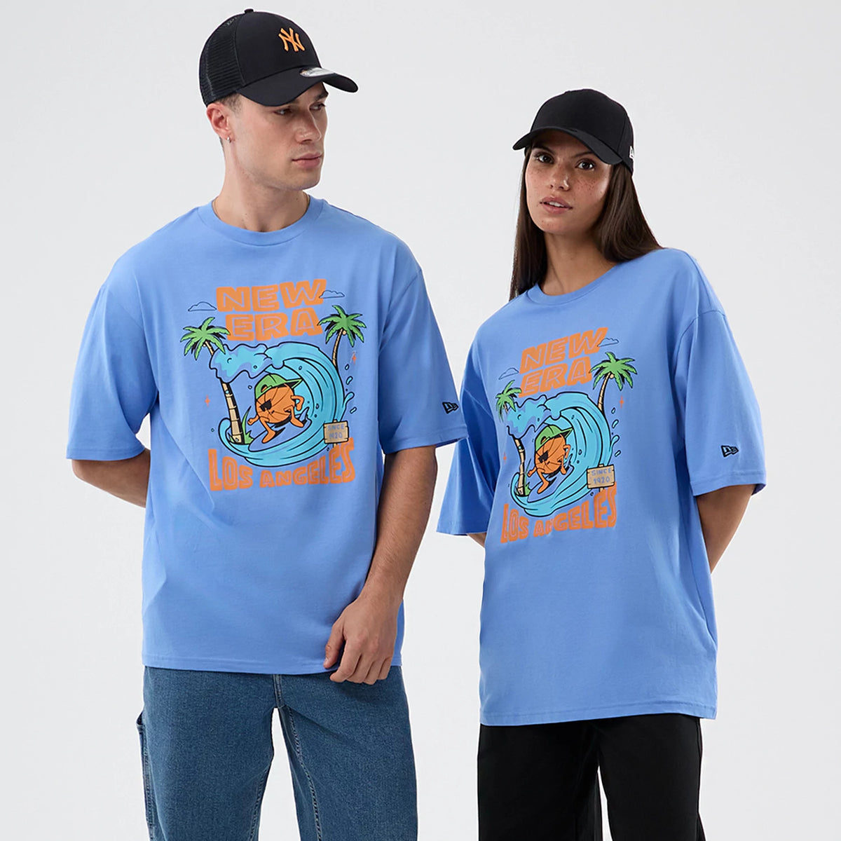 New Era Basketball Character Graphic Pastel Blue T-Shirt, front view on two models featuring a colourful cartoon graphic of a surfer in front of a wave with palm trees, prominent New Era and Los Angeles text.