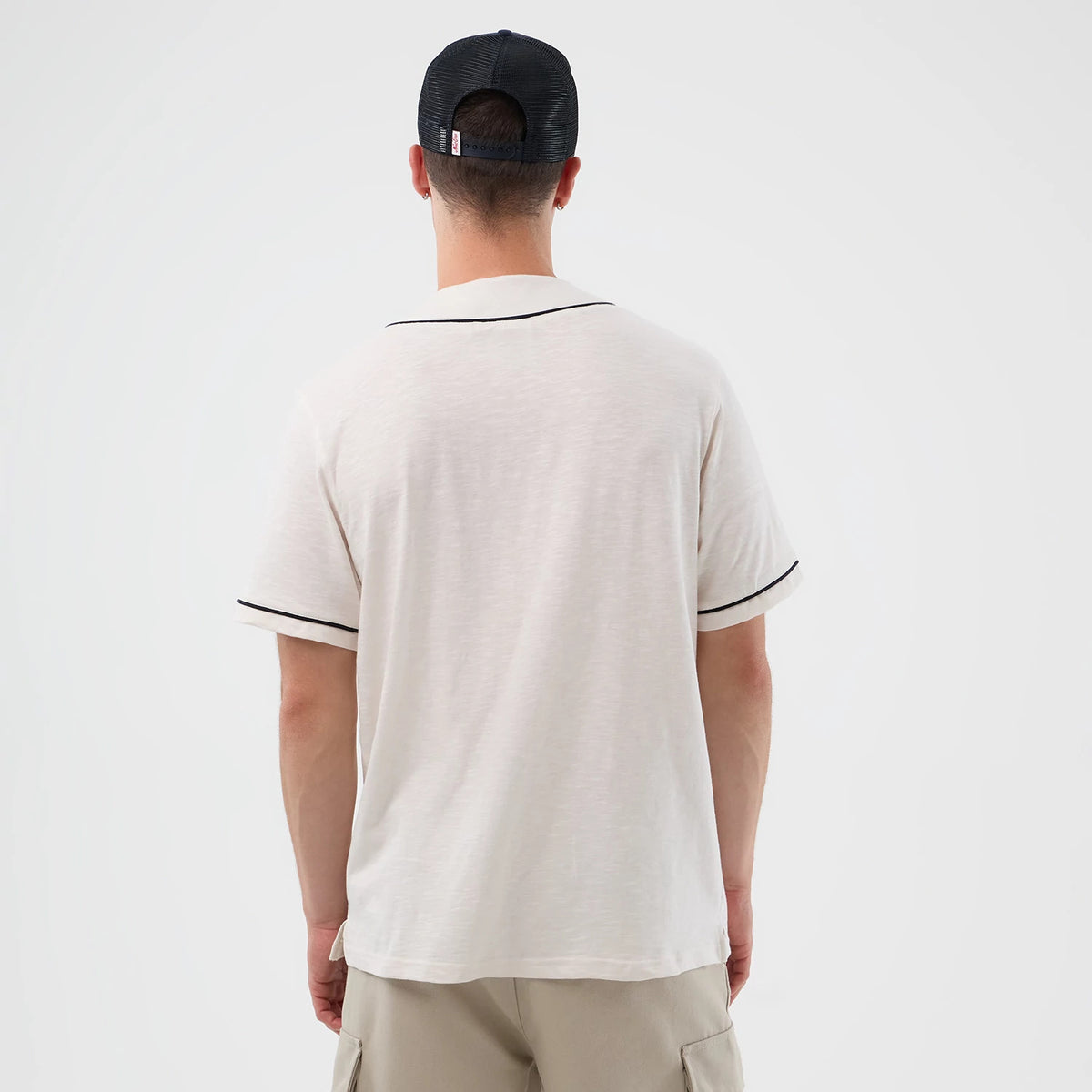 New Era Slub Open White Baseball Jersey, back view on a model wearing a black mesh-back cap, showcasing a plain white jersey with black trim on the sleeves and collar, paired with loose-fitting beige shorts.