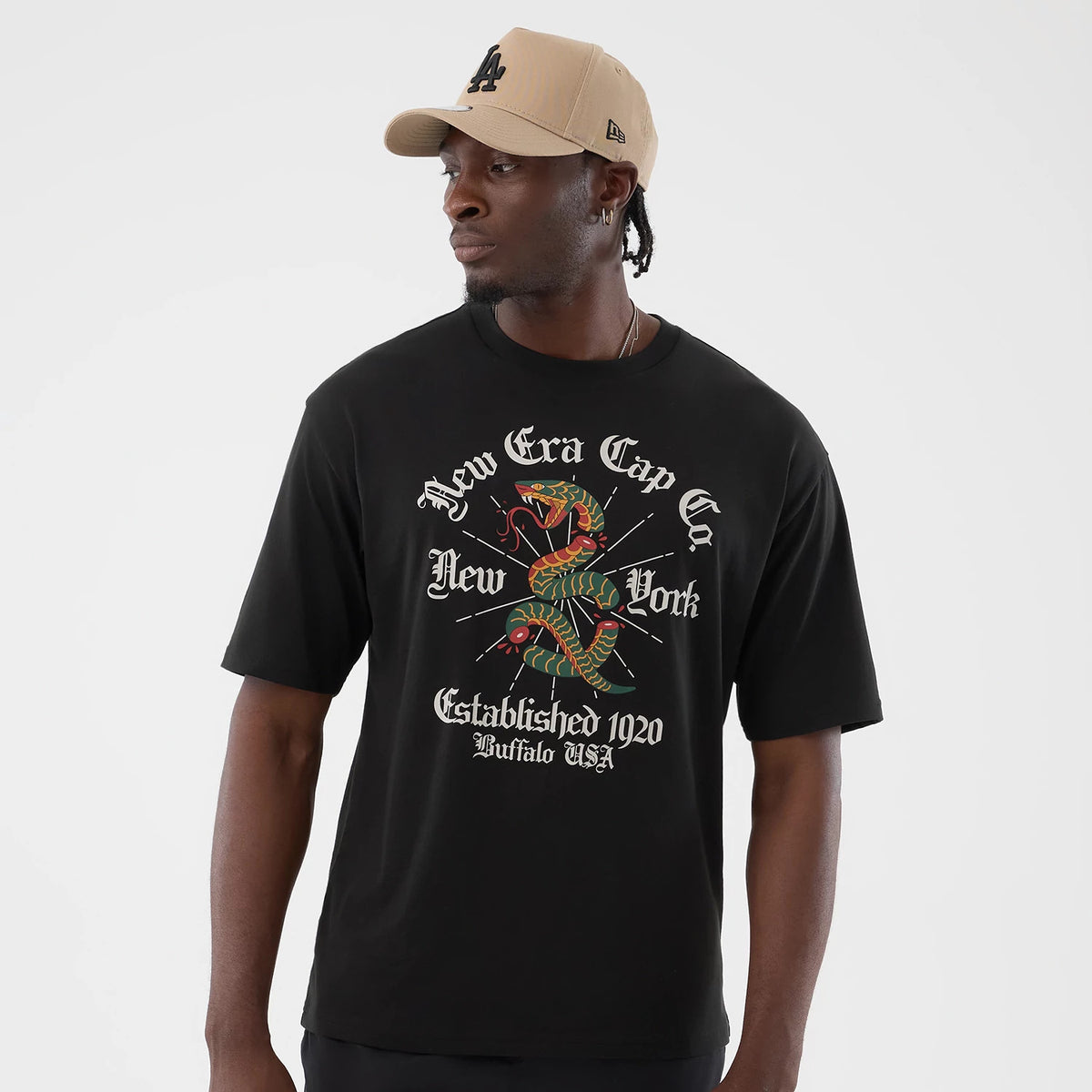 New Era Serpent Graphic Black Oversized T-Shirt, front view on a model wearing a beige cap, featuring a multicoloured serpent graphic in the centre, surrounded by white text with New Era Cap Co and Established 1920 Buffalo USA lettering.