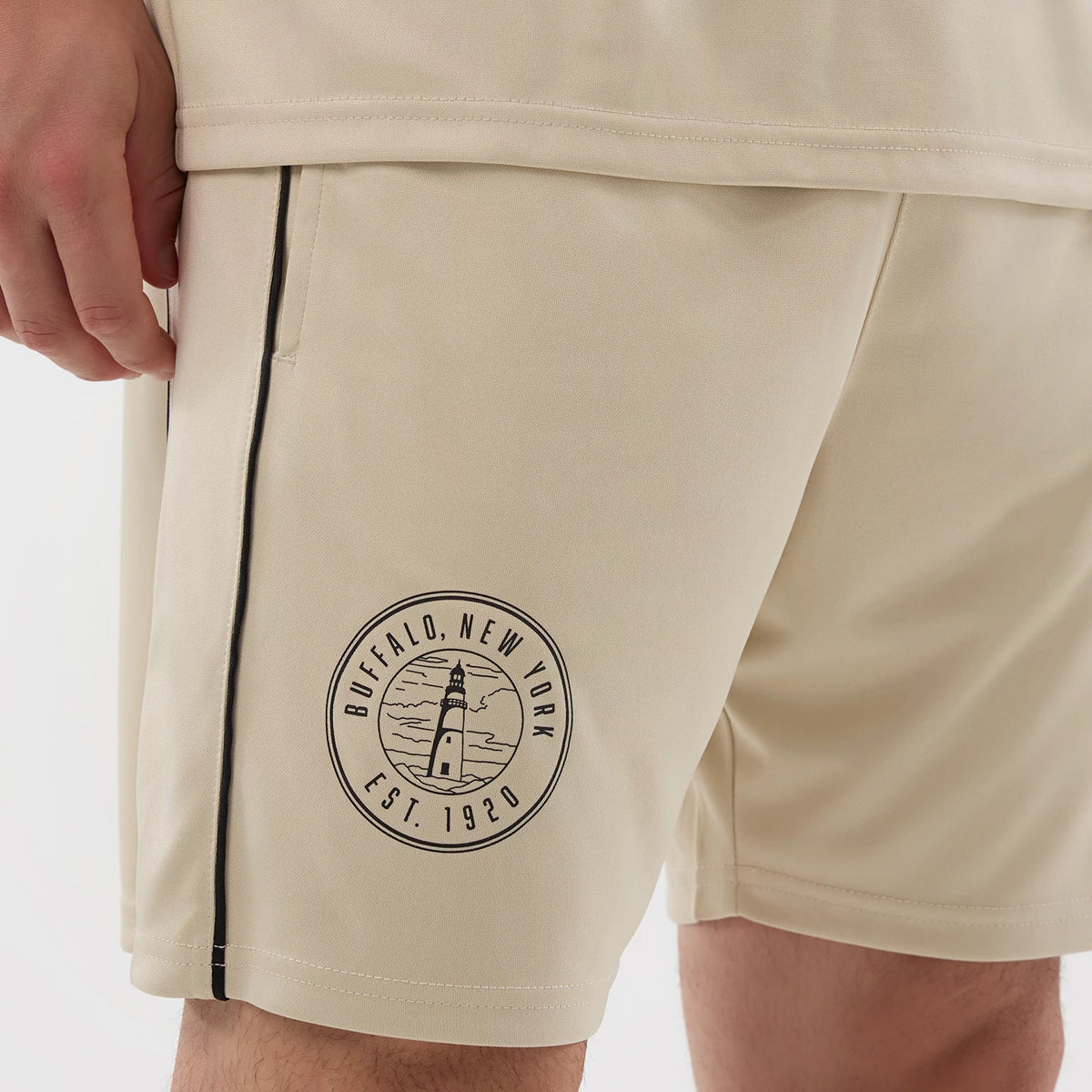 New Era Football Light Beige Shorts, front view showing black piping running down the sides, an embroidered circular Buffalo, New York emblem with a lighthouse graphic on the left leg, and a hidden side pocket beneath the waistband.