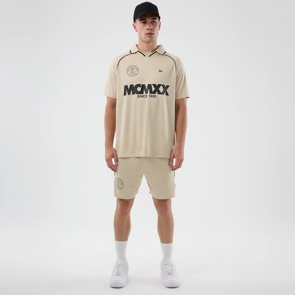 New Era Football Light Beige Shorts, front view on a model styled with a matching shirt and black cap, featuring an embroidered circular logo on the left leg, an elastic waistband, and a relaxed knee-length fit.