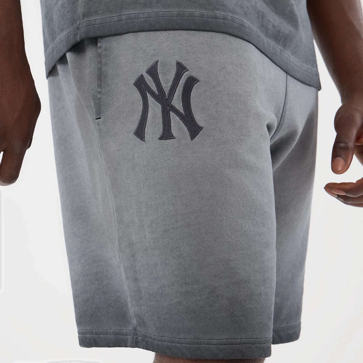 New Era New York Yankees MLB Washed Logo Black Shorts, side view on a model showing the dark grey washed fabric, a large black embroidered Yankees logo on the left leg, and a seam running along the hemline near the knee.