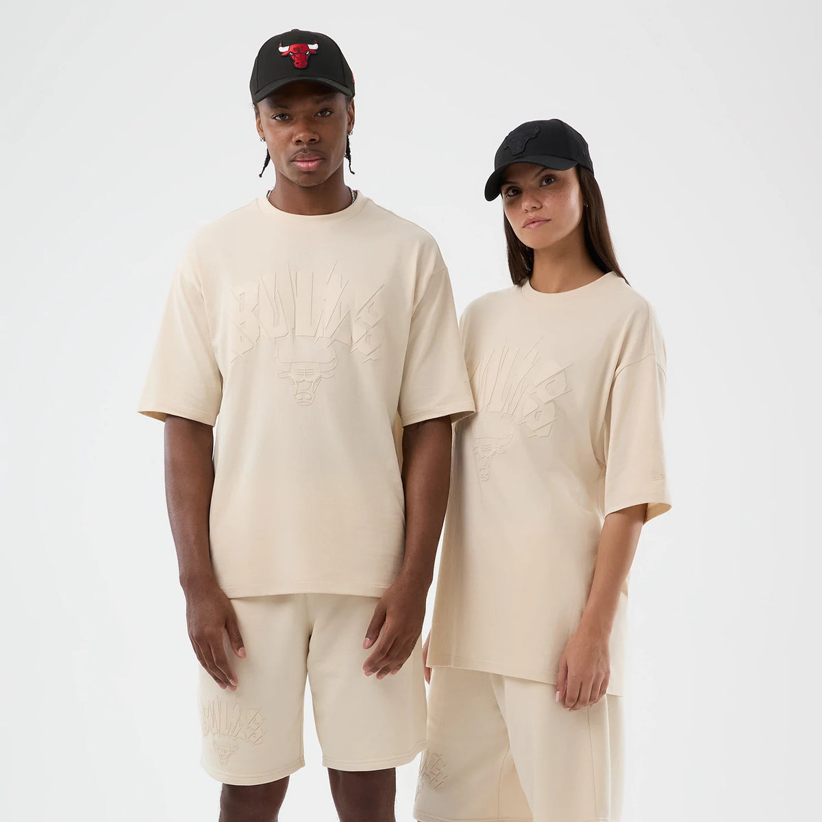 New Era Chicago Bulls NBA Core Plus Light Beige Oversized T-Shirt, front view on models with matching shorts and black caps, showing a raised Bulls logo and lettering across the chest and a embroidered New Era logo on the sleeve.