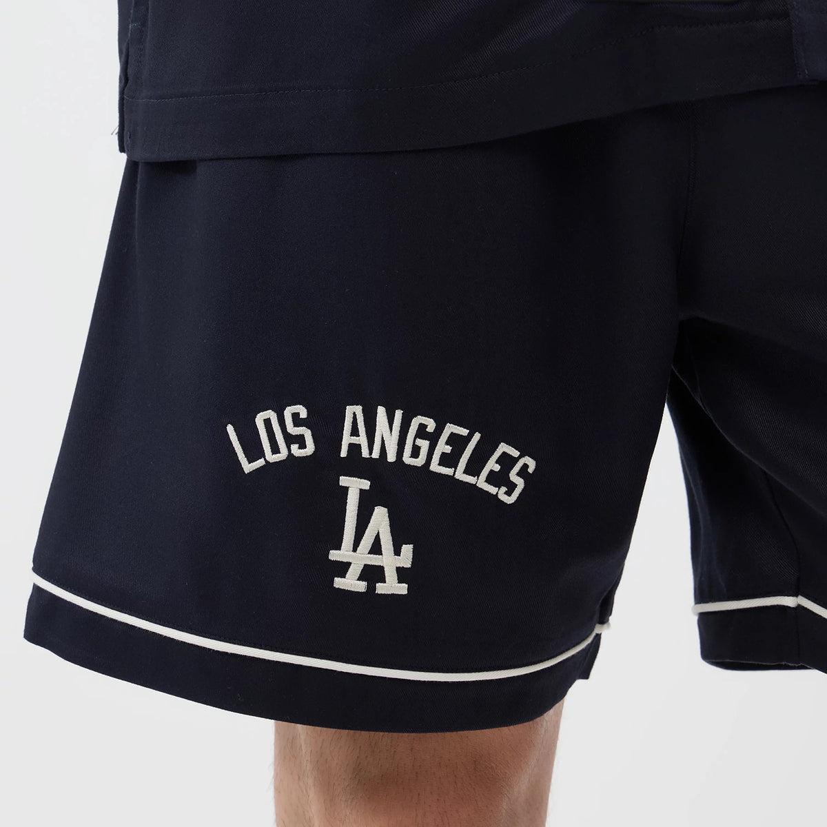 New Era LA Dodgers MLB Woven Navy Shorts, front view showing the embroidered Los Angeles Dodgers logo near the hem, with contrasting white piping along the edge and overlapping fabric detail at the top, styled with a matching jacket.