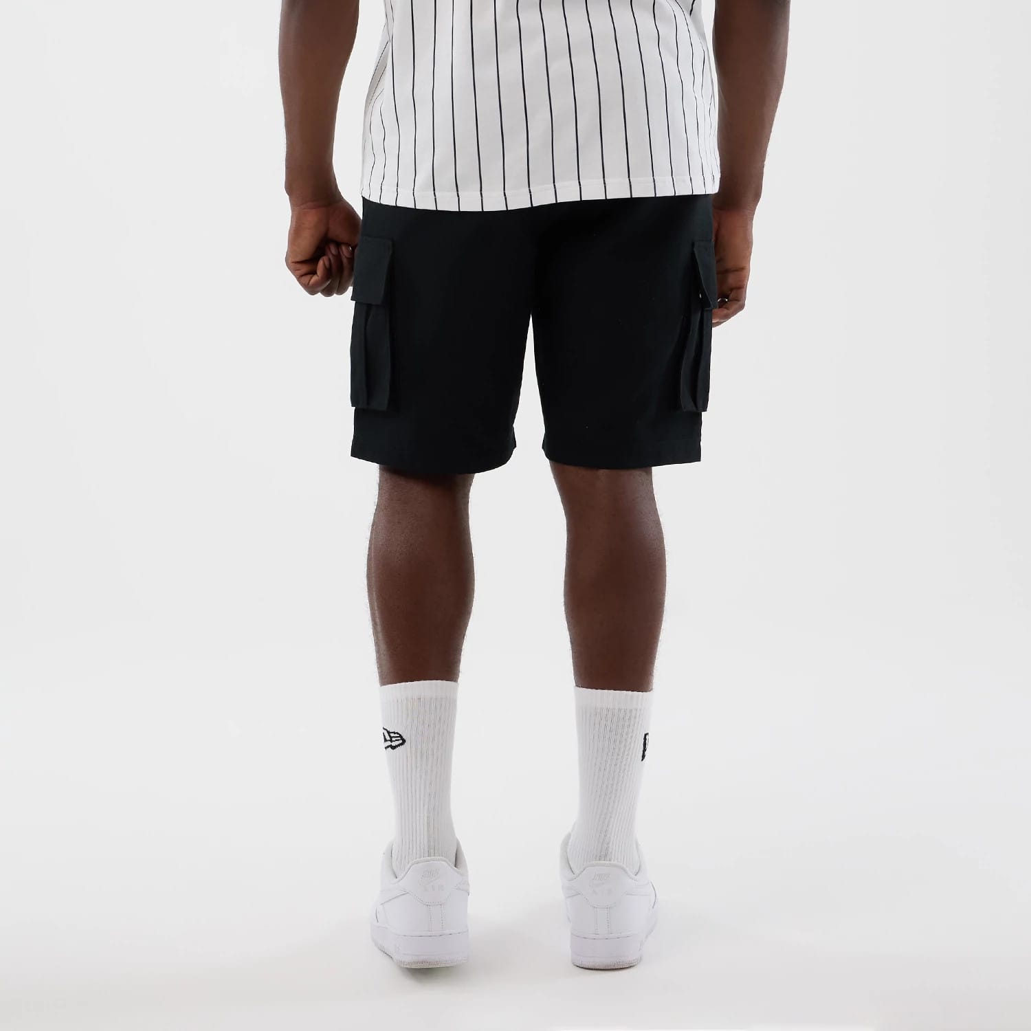 New Era Black Cargo Shorts, back view on a model featuring two large flap pockets on the sides of the shorts, paired with a white pinstripe top and white trainers, highlighting the casual and practical style of the shorts.