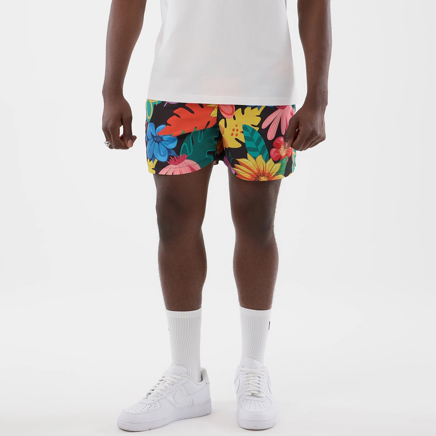 New Era Floral Black Swimming Shorts, front view on-model featuring a floral design with yellow, red, pink, blue, and green flowers, complemented by a plain white t-shirt, white socks, and matching white trainers.