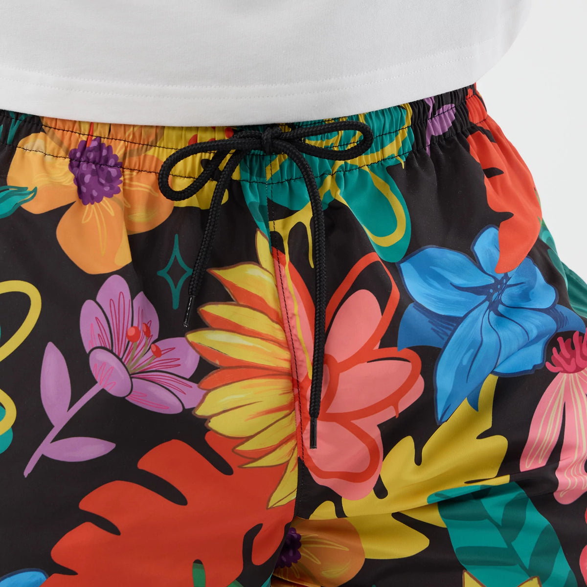 New Era Floral Black Swimming Shorts, front view showing a tropical floral print in pink, orange, blue, and green with an elastic waistband, black drawstring tied at the front, and a glimpse of a white top at the waistband's edge.