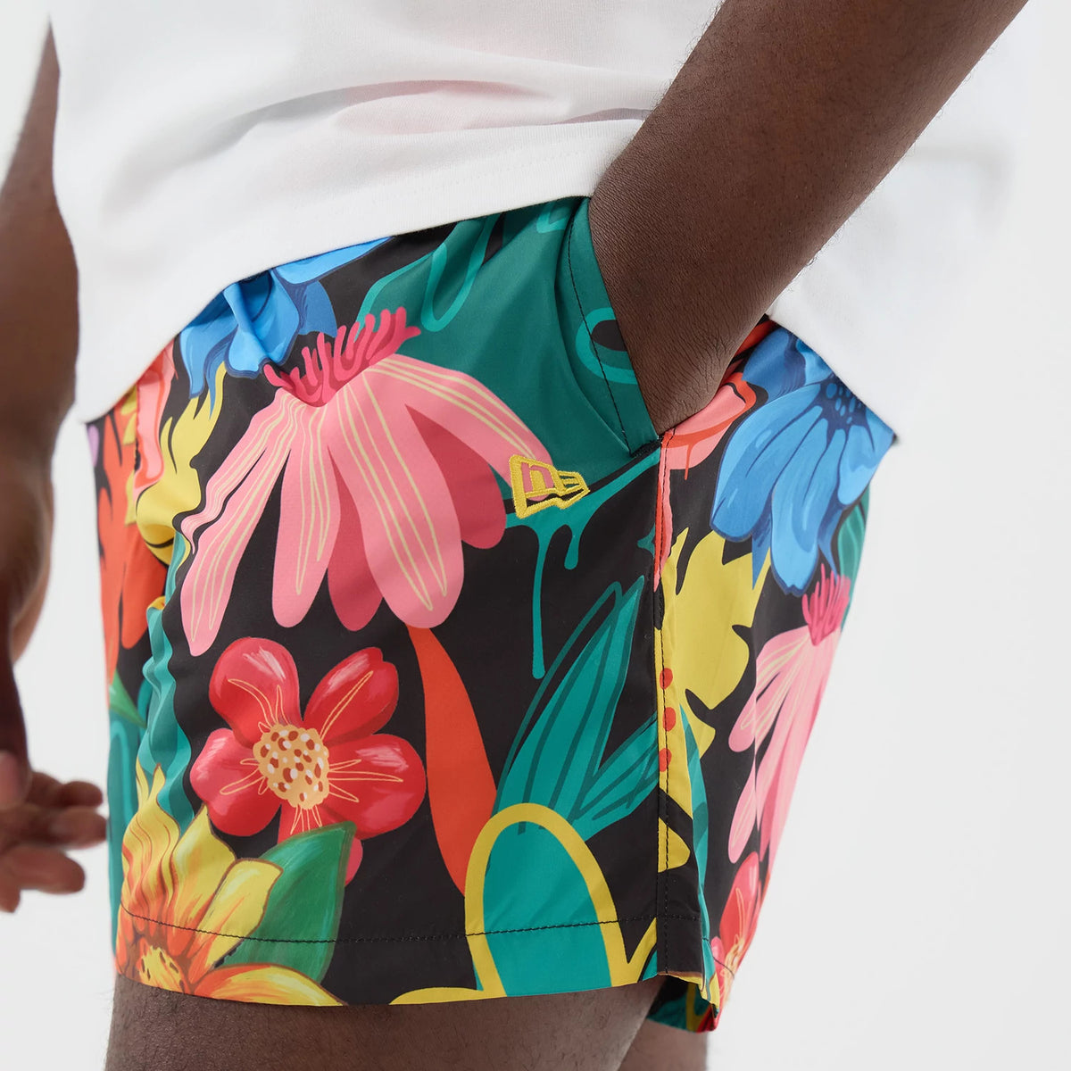 New Era Floral Black Swimming Shorts, side view on a model showing a floral pattern in pink, red, yellow, and blue, with a small raised New Era logo in yellow above the right hem, paired with a white top.