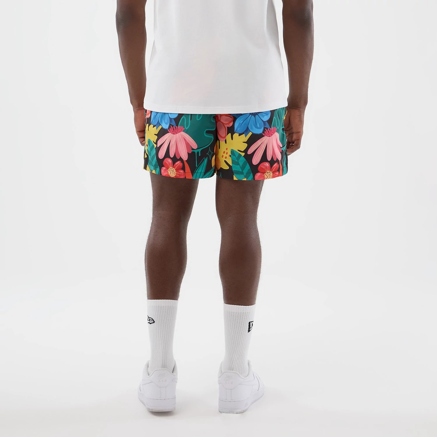 New Era Floral Black Swimming Shorts, back view on a model featuring a floral print with large pink, blue and red flowers on a black background, complemented by a plain white T-shirt, white socks, and white trainers.