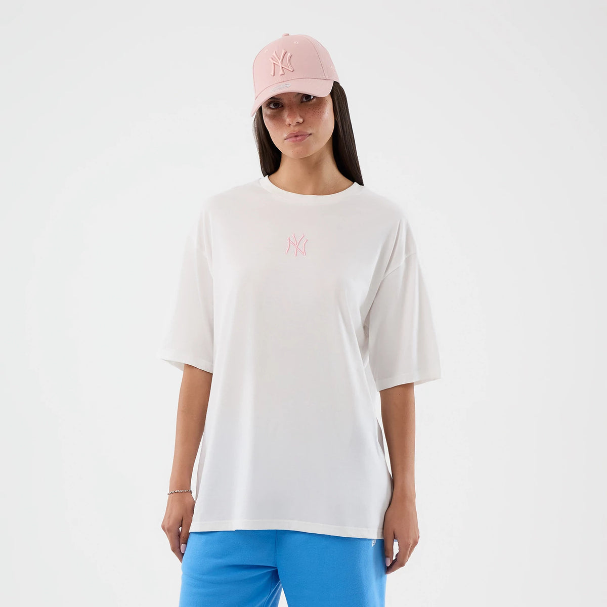 New Era New York Yankees MLB Midi League Essential White Oversized T-Shirt, front view on a model paired with a pink cap and blue bottoms, featuring a small pink embroidered Yankees logo centred on the chest of the loose-fitting white tee.