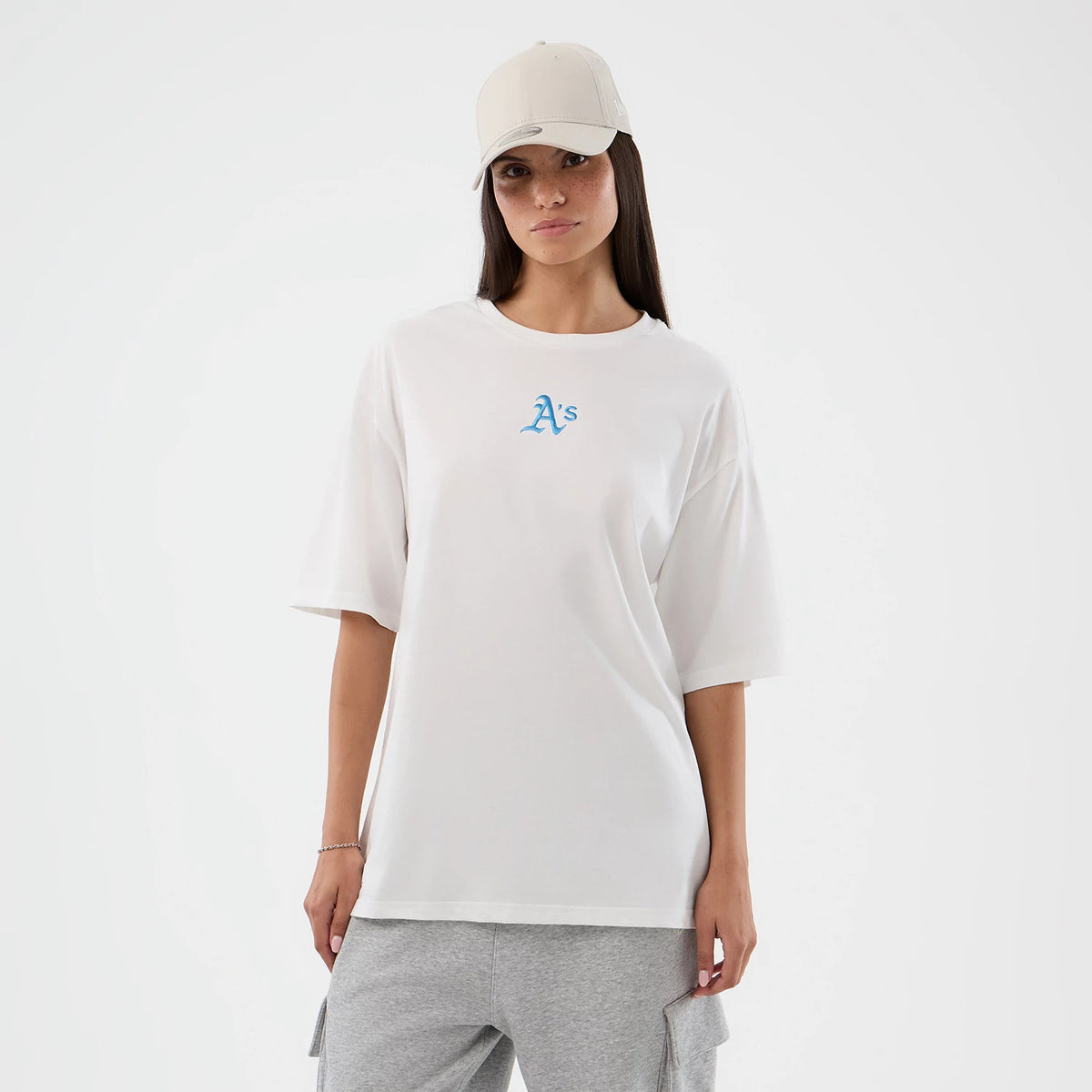 New Era Athletics MLB League Essential White Oversized T-Shirt, front view on-model showcasing a relaxed fit with a blue embroidered A's logo centred on the chest, short sleeves and styled with grey trousers and a neutral cap.