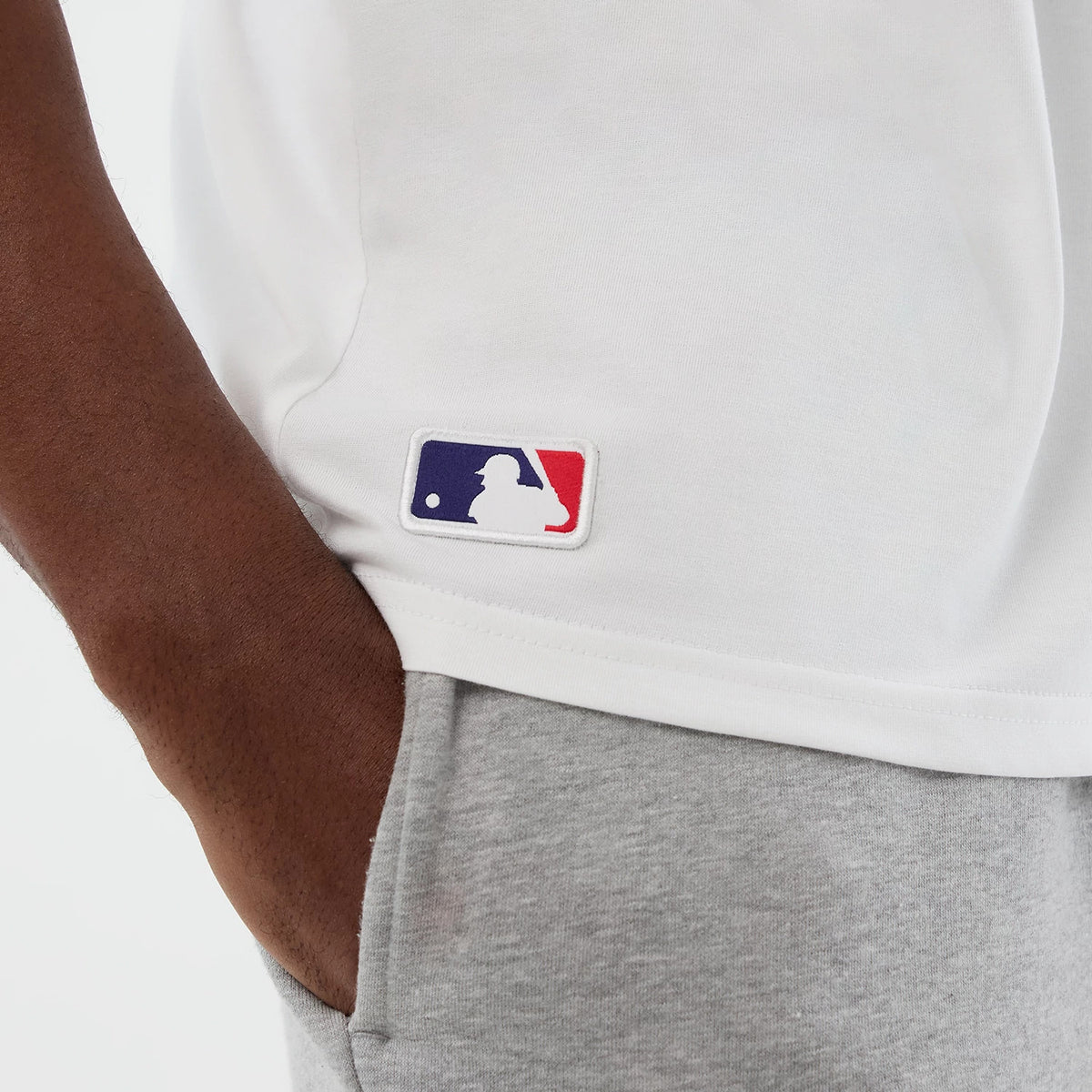 New Era Athletics MLB League Essential White Oversized T-Shirt, three-quarter view showing a model wearing grey joggers with a white embroidered MLB logo patch near the hem of the white t-shirt, providing a casual and relaxed styling.