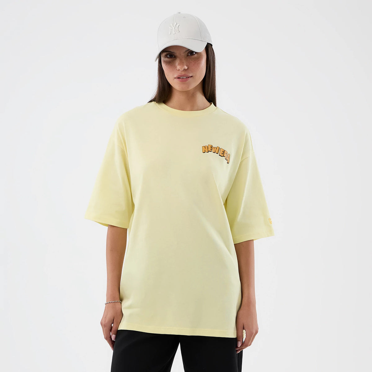 New Era Fun Beach Party Graphic Pastel Yellow Oversized T-Shirt, front view on a model, featuring the New Era lettering in orange printed on the left chest, short sleeves, and a relaxed fit with a plain front panel.