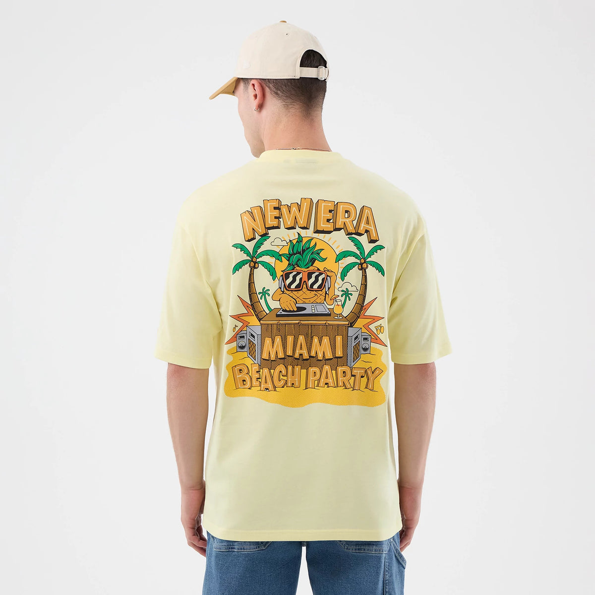 New Era Fun Beach Party Graphic Pastel Yellow Oversized T-Shirt, back view on a model wearing blue jeans and a cream cap, featuring an oversized graphic with palm trees, sun, and festive lettering reading New Era Miami Beach Party.