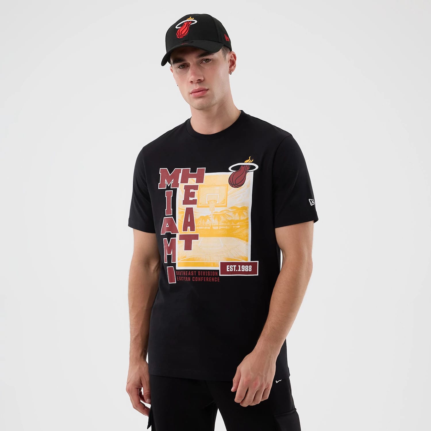 New Era Miami Heat NBA Graphic Black T-Shirt, front view on-model showing a graphic with Miami Heat text in red and yellow on the chest, a printed team logo on the upper right, and a New Era logo on the left sleeve.