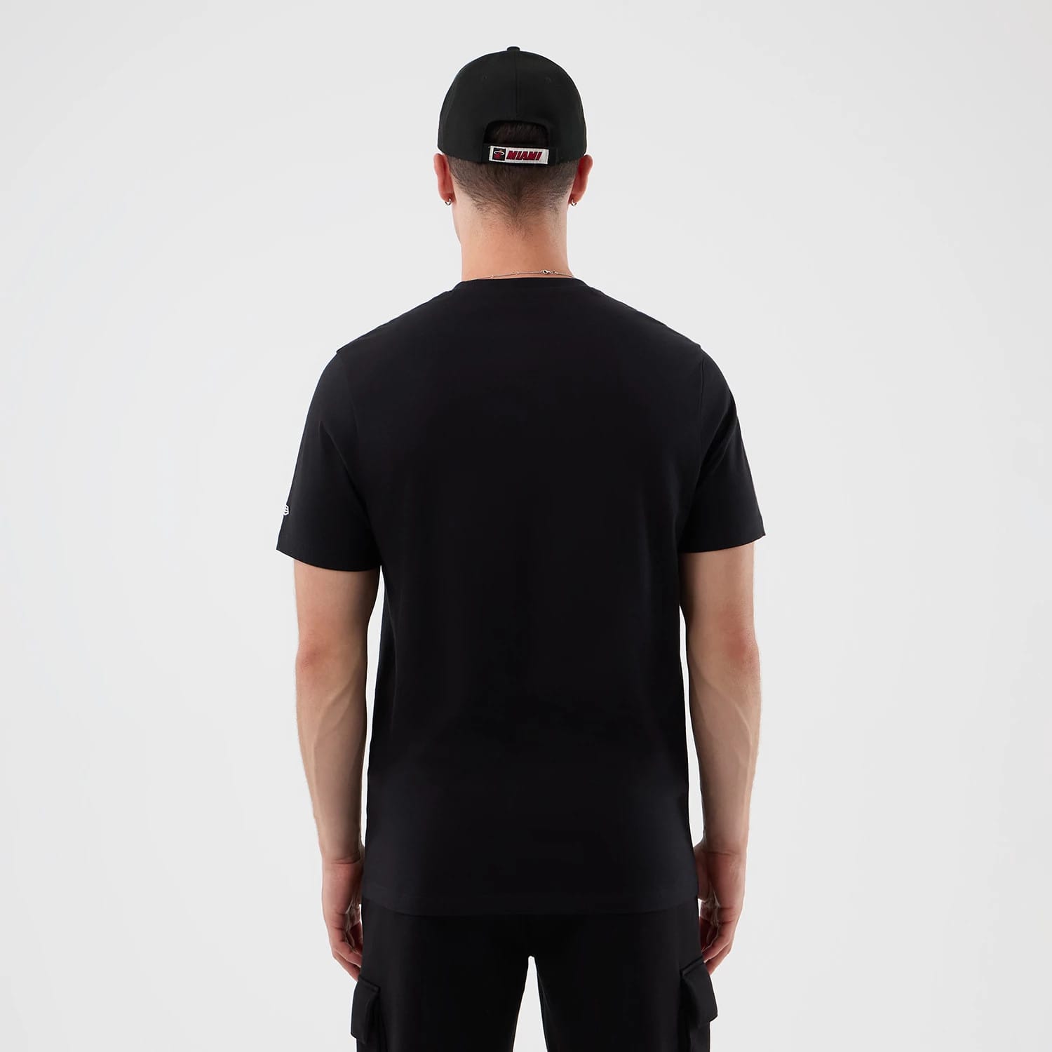 New Era Miami Heat NBA Graphic Black T-Shirt, back view on a model paired with black shorts and cap, showing a plain black back with short sleeves, a small logo embroidered on the left sleeve, and a tag on the back of the cap.
