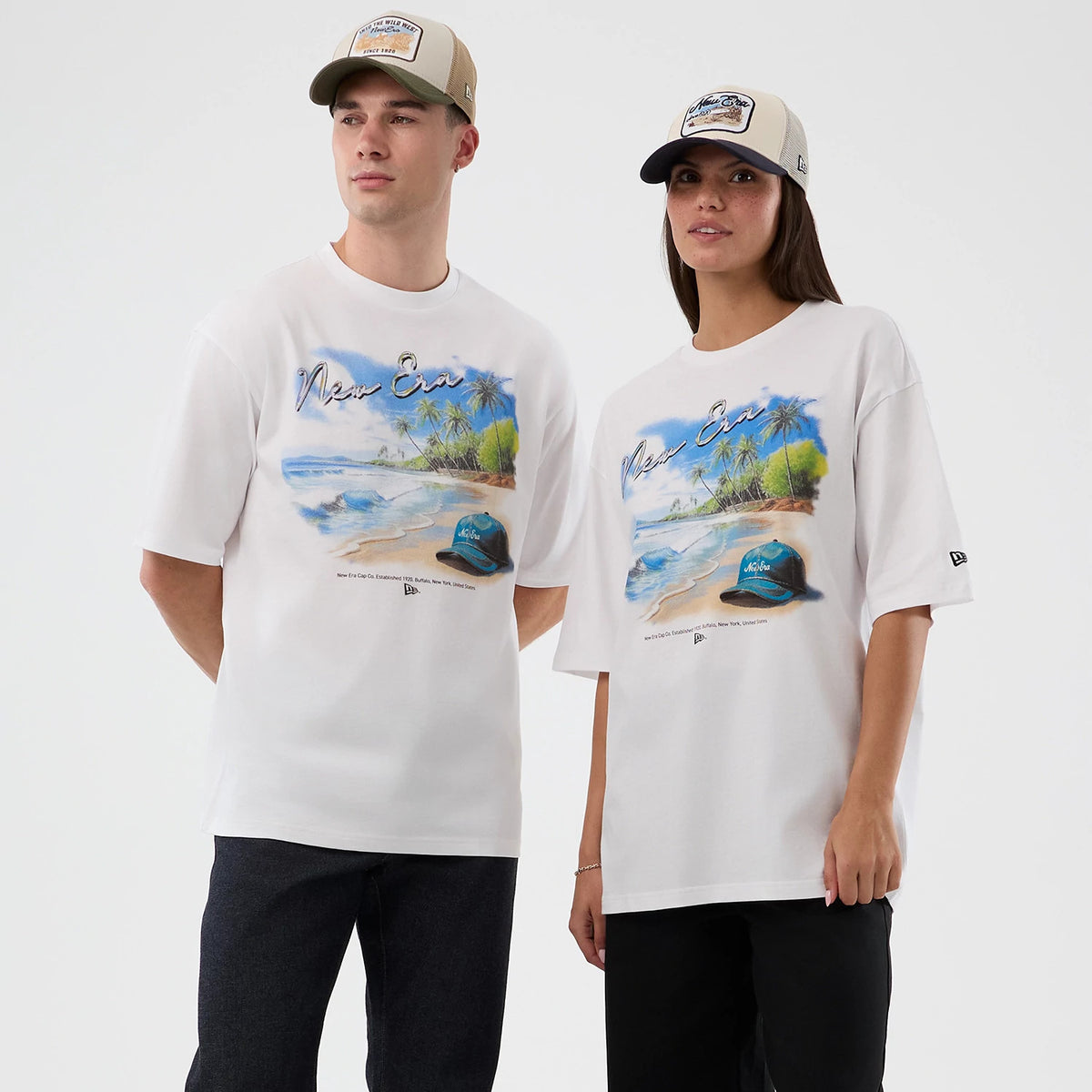 New Era Beach Graphic White Oversized T-Shirt, on-model view highlighting the beach scene print with palm trees and waves across the chest, a printed New Era logo on the left sleeve and worn with complementary caps.