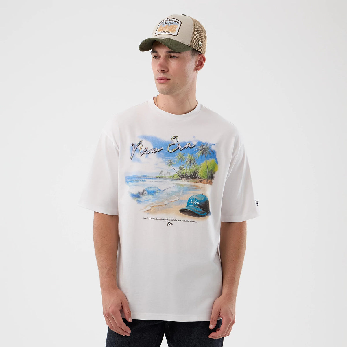 New Era Beach Graphic White Oversized T-Shirt, front view on-model showcasing a beach scene with palm trees and ocean waves on the chest, the New Era script above, and a small printed logo at the base near the hem on the left side.