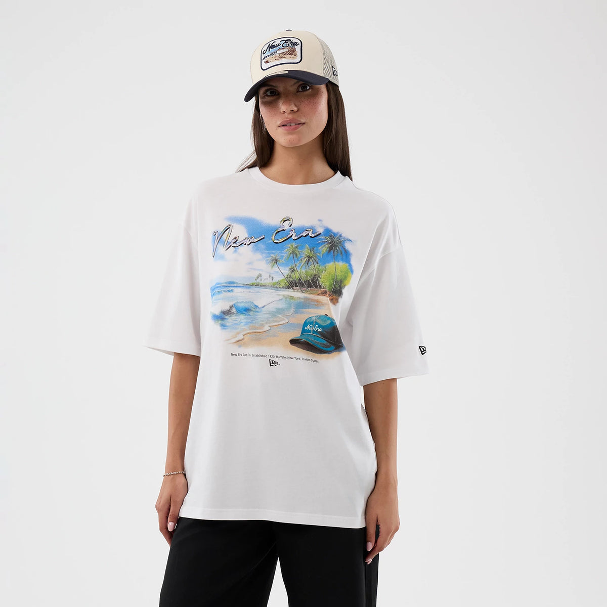New Era Beach Graphic White Oversized T-Shirt, front view on-model, featuring a large beach print with palm trees and waves, New Era script text across the chest, and a small embroidered logo near the bottom hem on a white oversized silhouette.