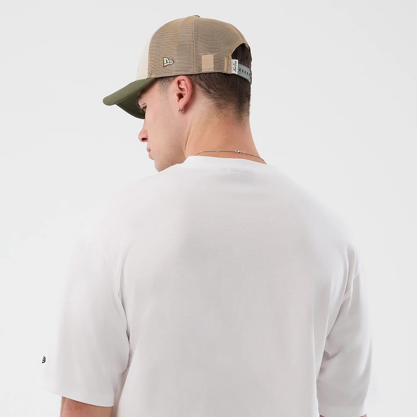 New Era Beach Graphic White Oversized T-Shirt, back view on-model showing the plain white back panel without graphics, a small embroidered New Era logo on the left sleeve, and a model wearing a tan mesh cap with an embroidered side logo.
