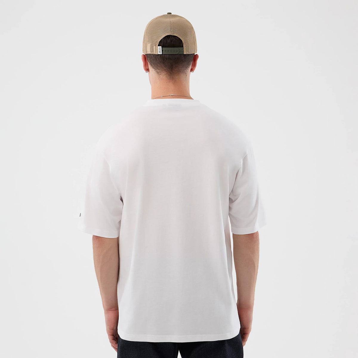 New Era Beach Graphic White Oversized T-Shirt, back view on a model with a light brown cap, displaying a plain white back with relaxed short sleeves and a New Era flag embroidered on the left sleeve near the cuff.