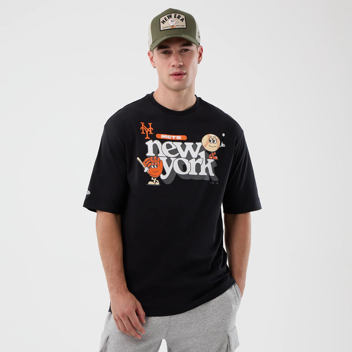 New Era New York Mets MLB Icon Graphic Black T-Shirt, front view on a model styled with a cap, featuring a large graphic design with cartoon characters and Mets New York text in grey and orange across the chest.
