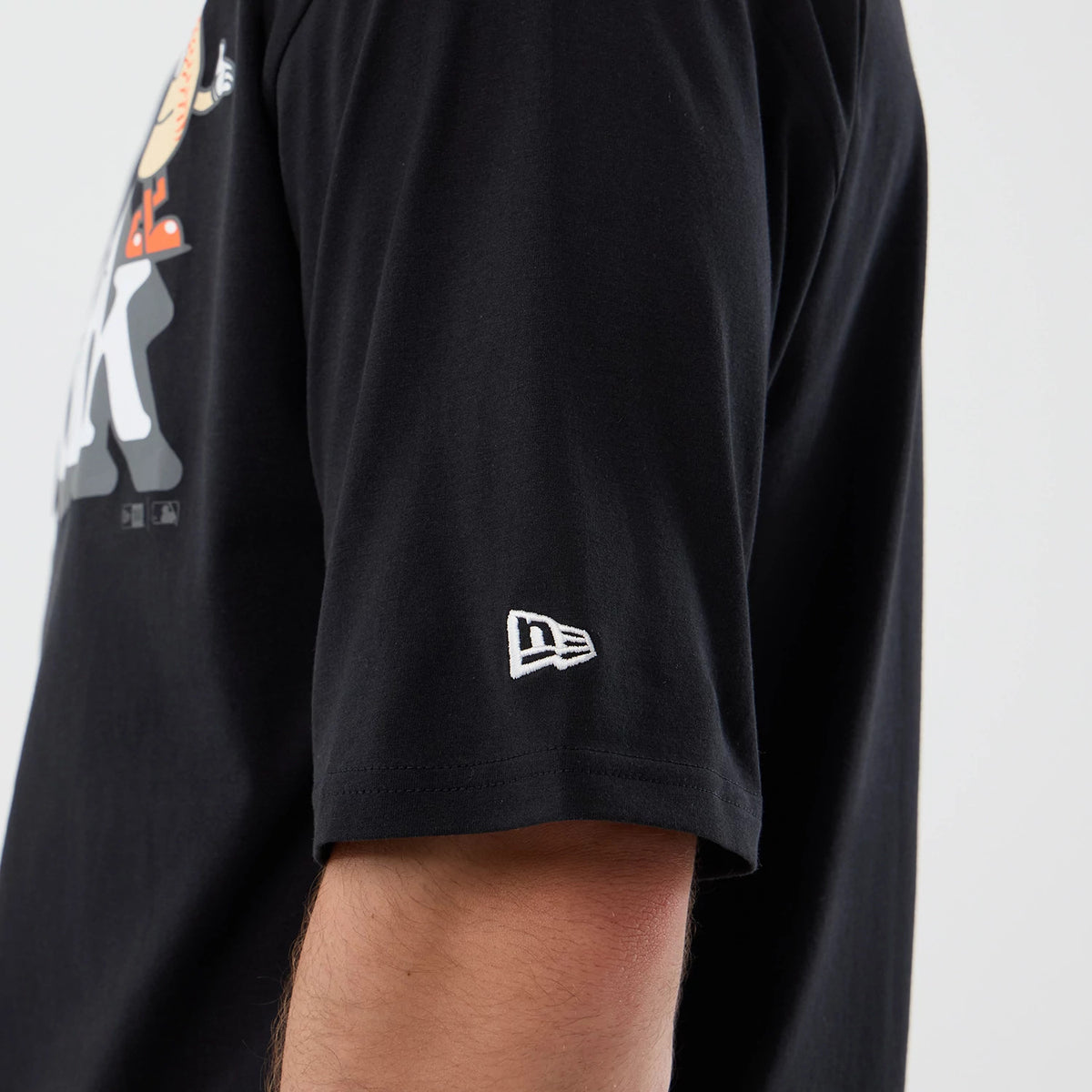 New Era New York Mets MLB Icon Graphic Black T-Shirt, side view on a model showing the embroidered white New Era logo on the left sleeve, a view of the Mets graphic on the chest, and the black fabric of the T-shirt.