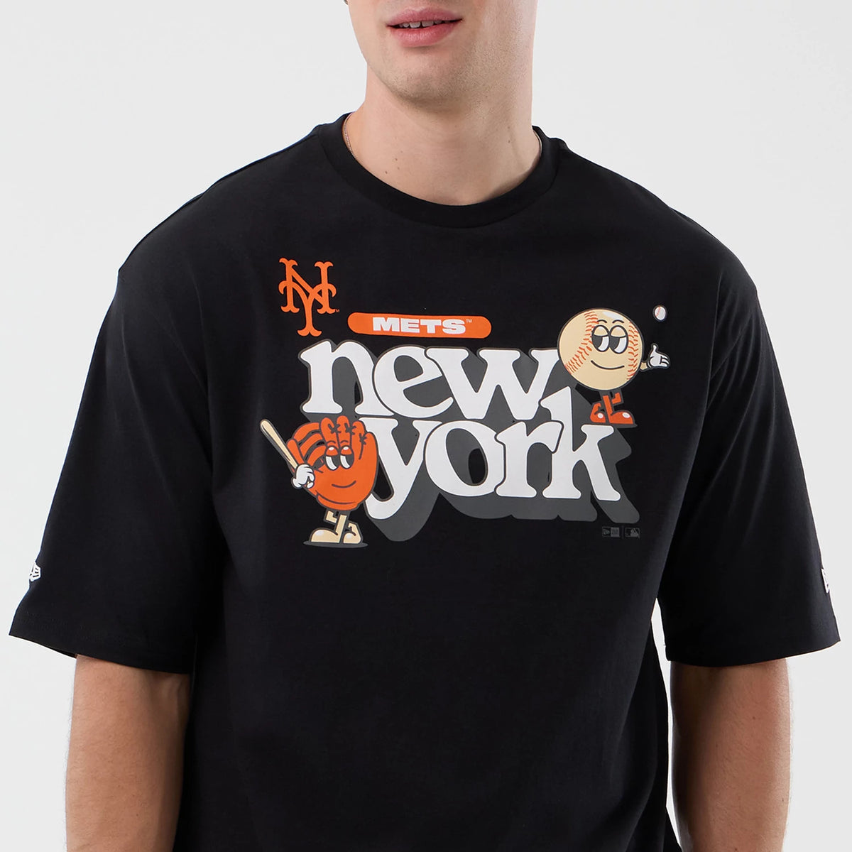 New Era New York Mets MLB Icon Graphic Black T-Shirt, front view on a model showing a colourful graphic with new york in large letters across the chest, an orange Mets logo above, and cartoon characters including a baseball and glove on each side.