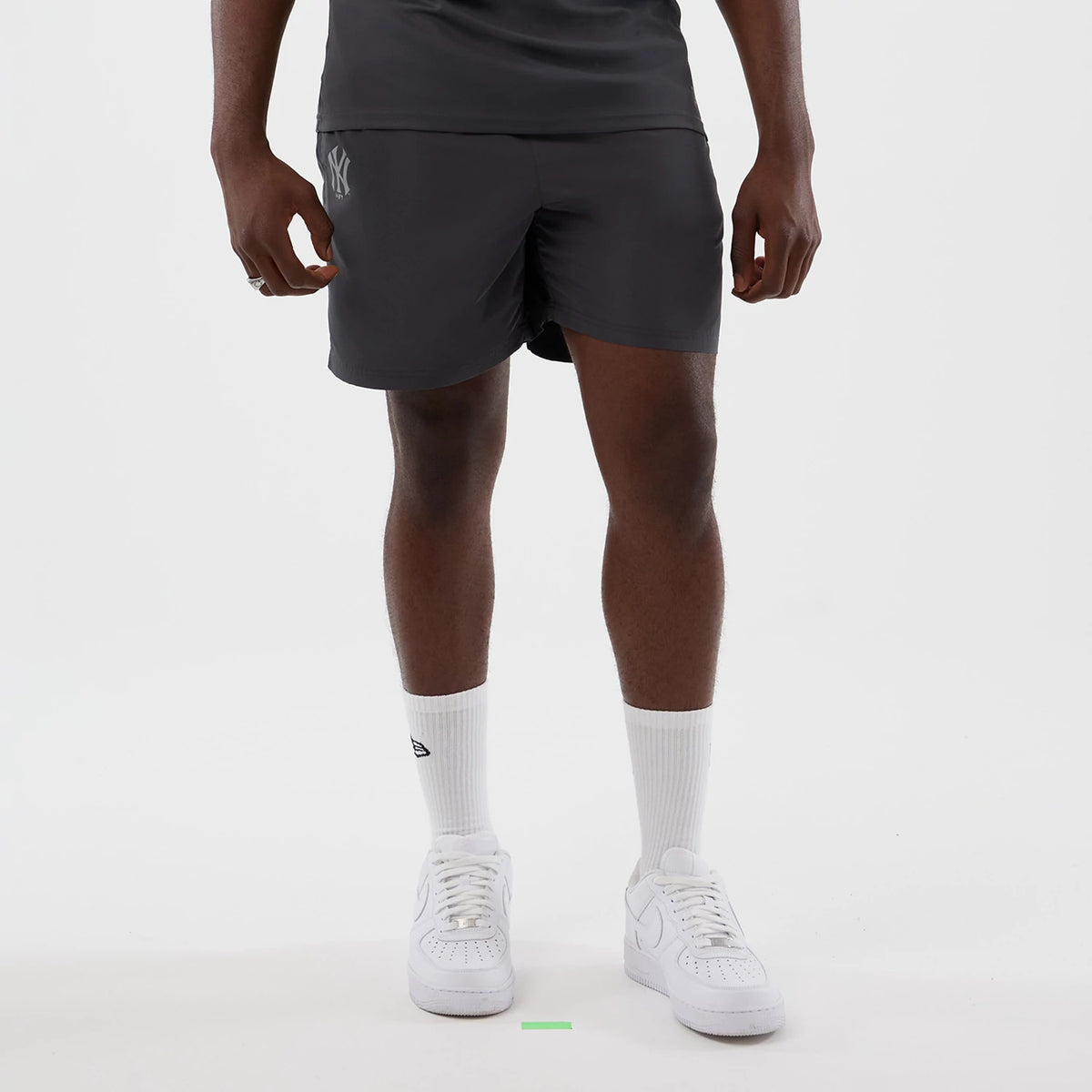 New Era New York Yankees MLB Core Plus Dark Grey Shorts, front view on-model showing the New York Yankees logo embroidered in light grey on the left leg, styled with a dark top, white socks with a small logo, and white trainers.