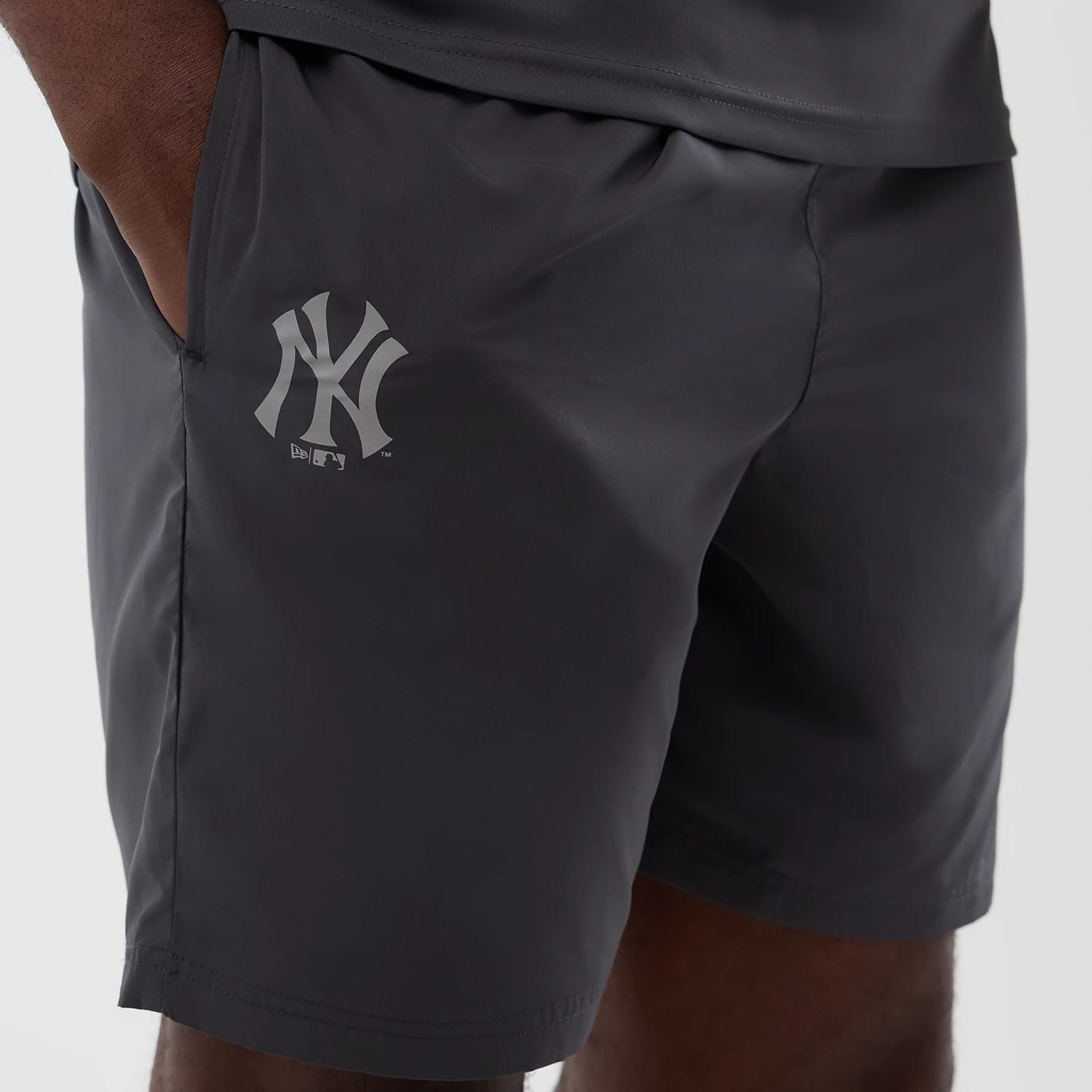 New Era New York Yankees MLB Core Plus Dark Grey Shorts, front view on a model showing the dark grey fabric with pockets, featuring a white printed NY Yankees logo on the left leg, and stitching along the seams for a finish.
